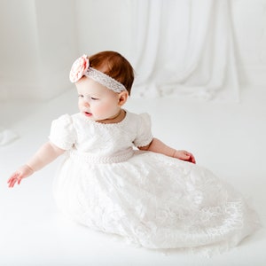 Puede incluir: Una ni&ntilde;a peque&ntilde;a con un vestido blanco de encaje y una diadema de flores rosa. El vestido tiene una cinta de sat&eacute;n alrededor de la cintura.