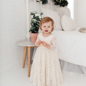 May include: A young girl wearing a white lace dress with long sleeves stands in front of a white bed with a white headboard and a green garland. The girl is looking at the camera and smiling.