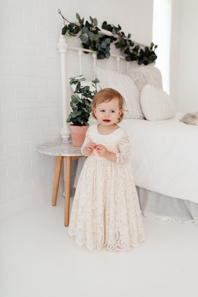 May include: A young girl wearing a white lace dress with long sleeves stands in front of a white bed with a white headboard and a green garland. The girl is looking at the camera and smiling.
