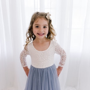 May include: A young girl wearing a white lace long-sleeved dress with a light blue tulle skirt. She is smiling and has a small bow in her hair.