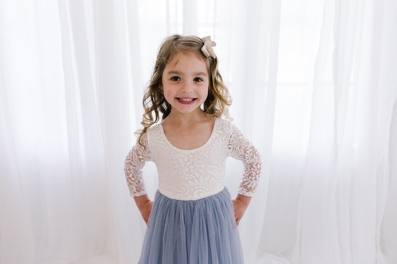 May include: A young girl wearing a white lace long-sleeved dress with a light blue tulle skirt. She is smiling and has a small bow in her hair.