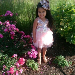 May include: A young girl wearing a pink and gold sequined dress with a tulle skirt and a large gold bow in her hair. She is standing in a garden with pink flowers.