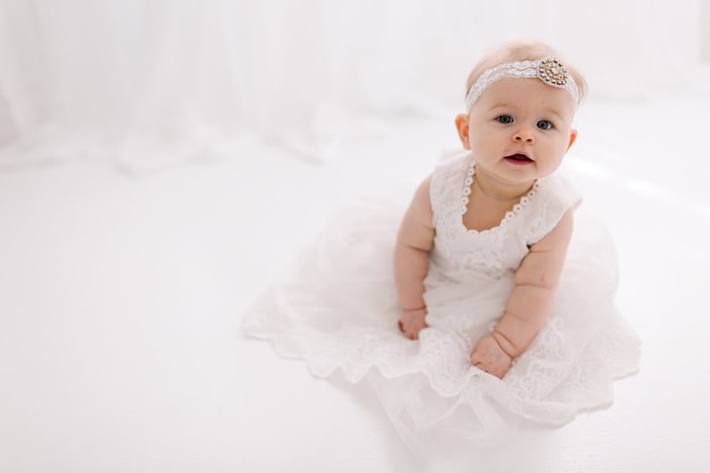 May include: A baby girl wearing a white lace dress and a headband with a jeweled accent. She is sitting on a white background and looking at the camera.