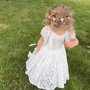 May include: A young girl wearing a white lace dress with a flower crown on her head. She is standing in a grassy field.
