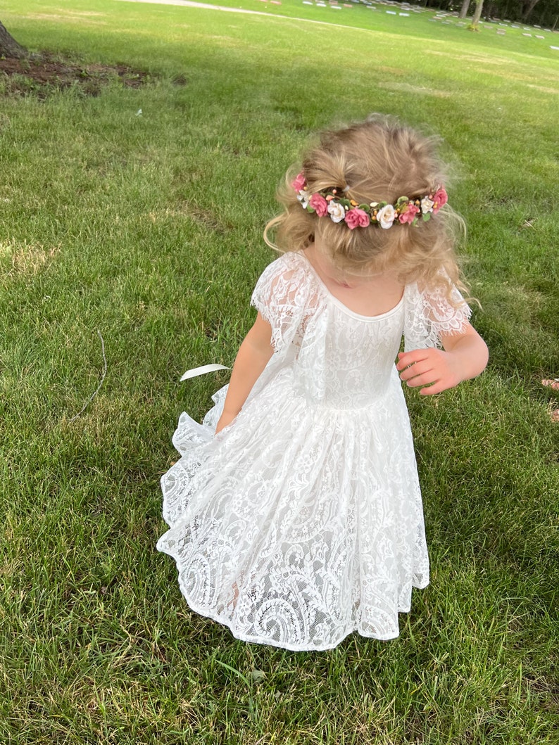 May include: A young girl wearing a white lace dress with a flower crown on her head. She is standing in a grassy field.