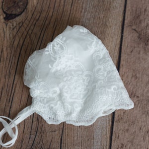 May include: White lace baby bonnet with a delicate floral pattern and a white satin ribbon tie.