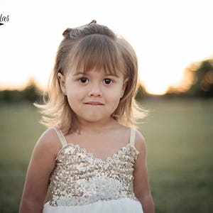 May include: A sleeveless dress featuring a bodice covered in shimmering gold sequins with a scalloped neckline edge. The dress has thin, light-colored straps and a plain white skirt portion visible at the bottom, worn by a young child with light brown hair.