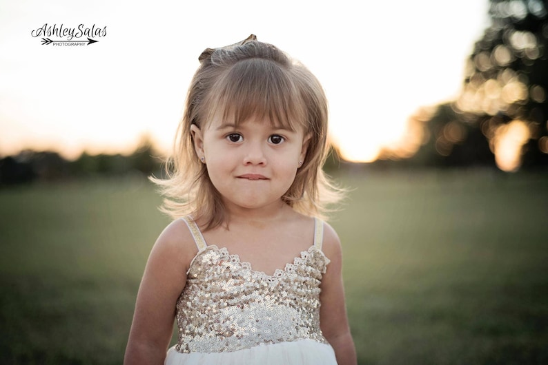 May include: A sleeveless dress featuring a bodice covered in shimmering gold sequins with a scalloped neckline edge. The dress has thin, light-colored straps and a plain white skirt portion visible at the bottom, worn by a young child with light brown hair.