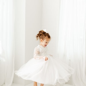May include: A young girl in a white tulle dress with lace sleeves twirls in a white room. She has curly brown hair and is wearing a white bow.