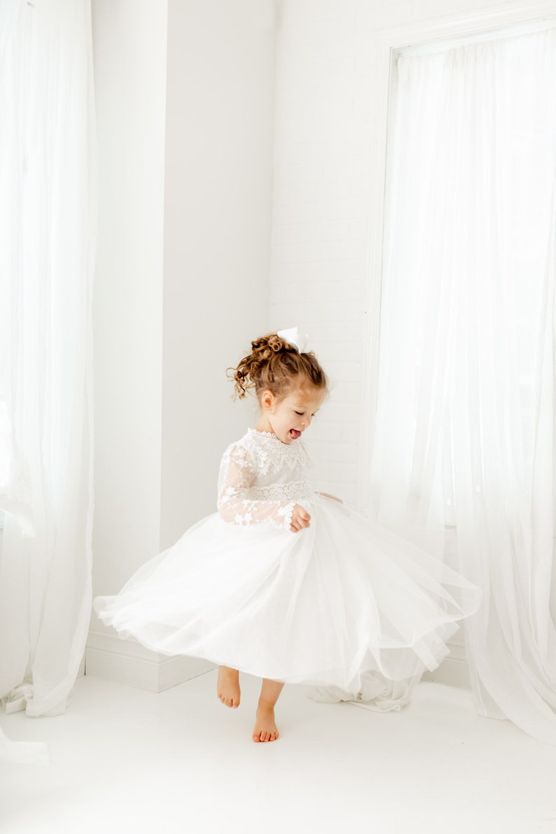 May include: A young girl in a white tulle dress with lace sleeves twirls in a white room. She has curly brown hair and is wearing a white bow.