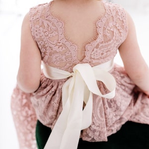 Pu&ograve; includere: Un abito in pizzo rosa con un nastro di raso bianco legato in un fiocco sul retro. L'abito ha una scollatura a festoni e una gonna ampia.