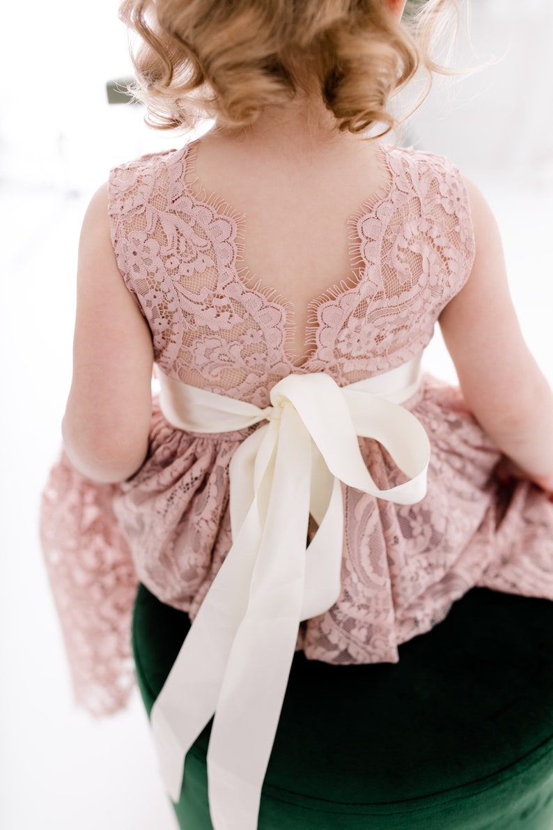 Pu&ograve; includere: Un abito in pizzo rosa con un nastro di raso bianco legato in un fiocco sul retro. L'abito ha una scollatura a festoni e una gonna ampia.