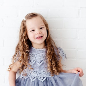 K&ouml;nnte beinhalten: Ein junges M&auml;dchen mit langen, lockigen Haaren l&auml;chelt, w&auml;hrend es ein hellblaues T&uuml;llkleid mit einer wei&szlig;en Spitzen&uuml;berlage tr&auml;gt. Das Kleid hat einen runden Ausschnitt und kurze &Auml;rmel.