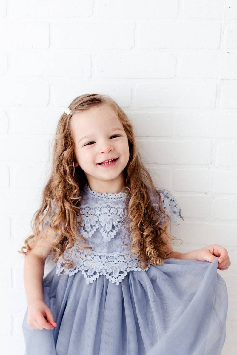 K&ouml;nnte beinhalten: Ein junges M&auml;dchen mit langen, lockigen Haaren l&auml;chelt, w&auml;hrend es ein hellblaues T&uuml;llkleid mit einer wei&szlig;en Spitzen&uuml;berlage tr&auml;gt. Das Kleid hat einen runden Ausschnitt und kurze &Auml;rmel.