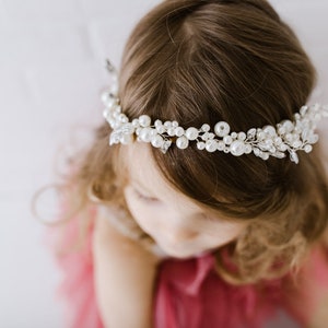May include: A young girl wearing a white pearl and crystal hair crown. The crown is made of delicate silver wire and features a cluster of pearls and crystals.