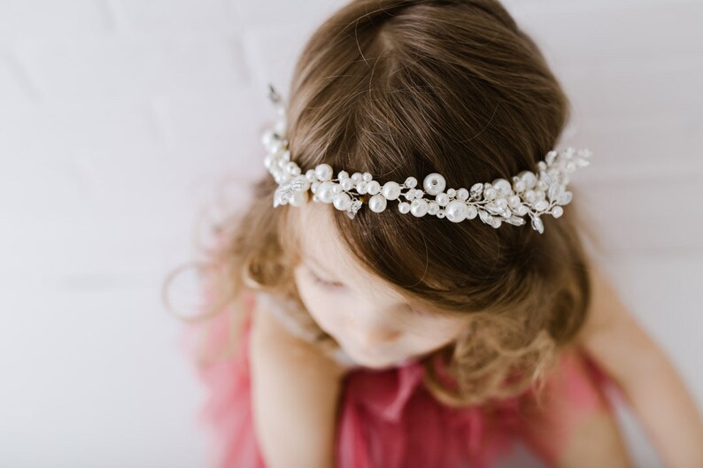 May include: A young girl wearing a white pearl and crystal hair crown. The crown is made of delicate silver wire and features a cluster of pearls and crystals.