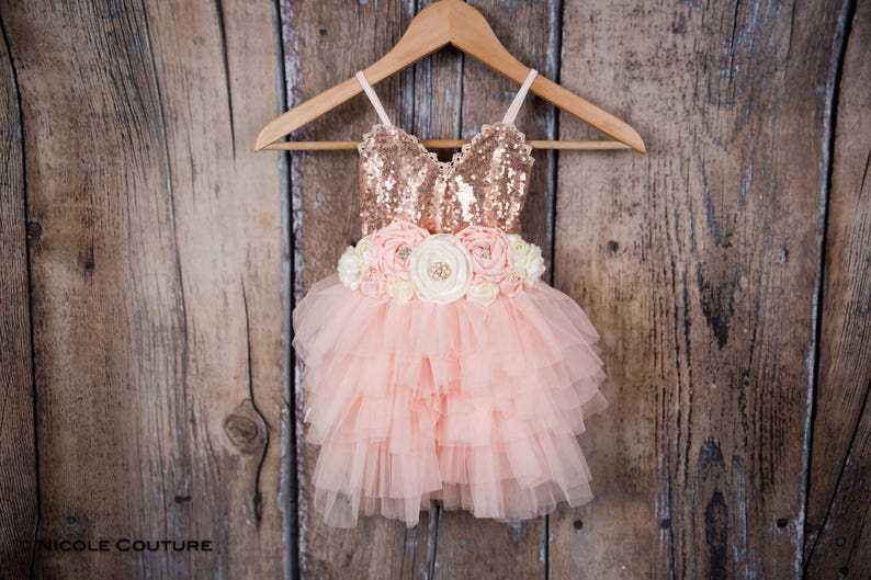 May include: A pink tulle dress with a rose gold sequin bodice and a flower sash. The dress is on a wooden hanger.