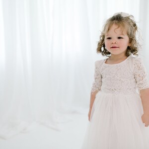 May include: A young girl wearing a white lace dress with a tulle skirt. She has curly blonde hair and is looking to the side.