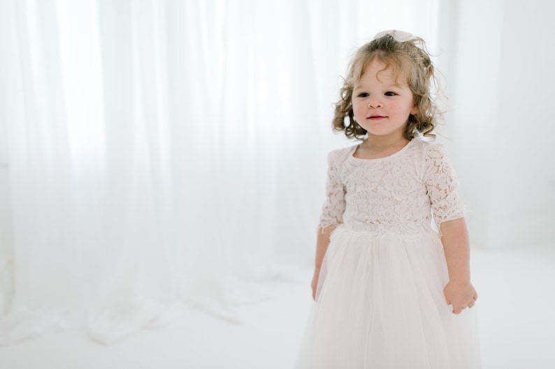 May include: A young girl wearing a white lace dress with a tulle skirt. She has curly blonde hair and is looking to the side.