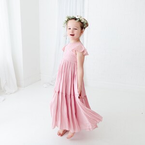 May include: A young girl wearing a pink, tiered maxi dress with a floral crown. The dress has a smocked bodice and short, ruffled sleeves.