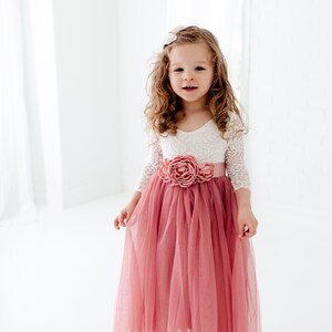 May include: A young girl wearing a white lace top with a pink tulle skirt and a pink flower sash. The skirt is long and flowing, and the girl is smiling.