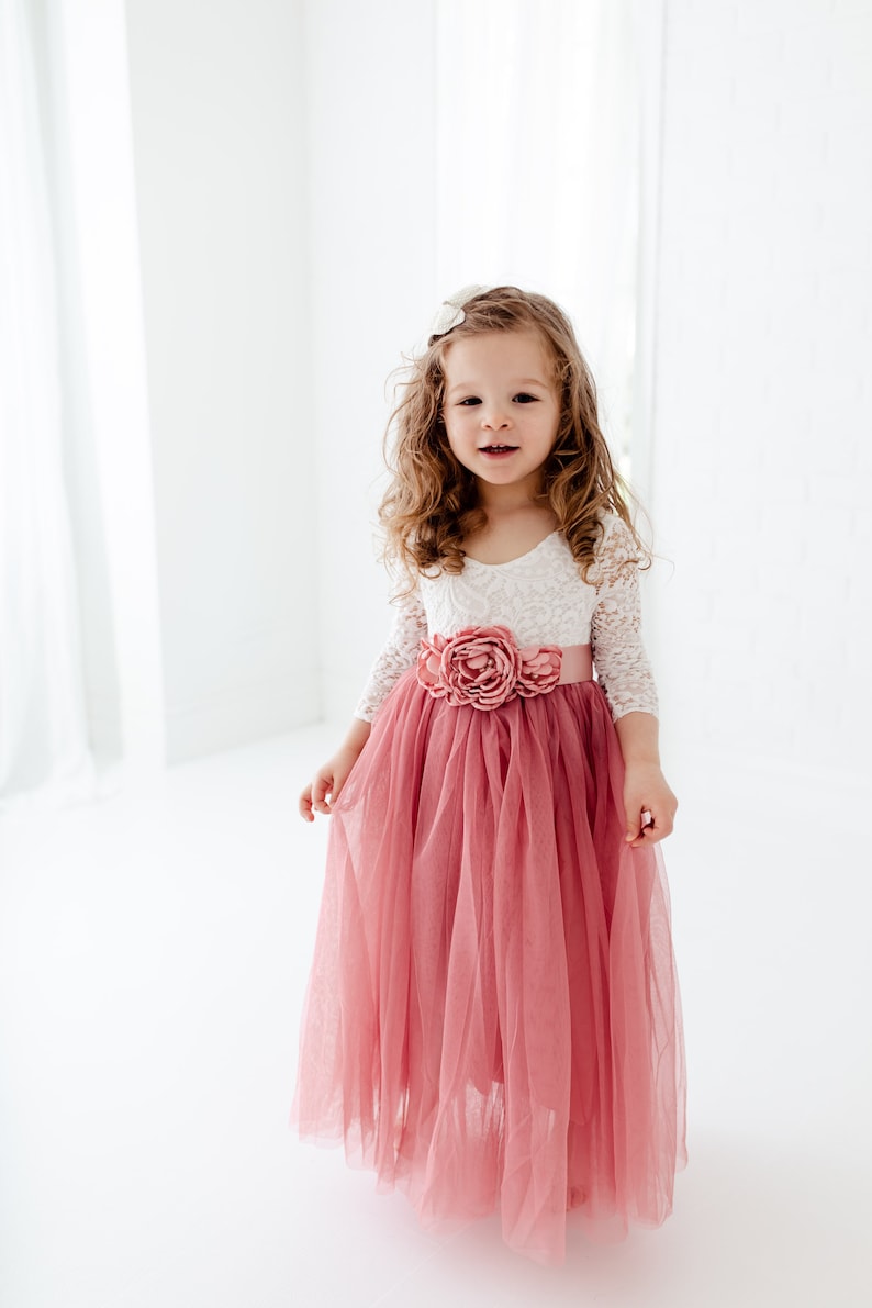 May include: A young girl wearing a white lace top with a pink tulle skirt and a pink flower sash. The skirt is long and flowing, and the girl is smiling.