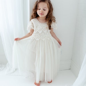 May include: A cream-colored girl's dress with a lace bodice and a flowing tulle skirt. The dress features short sleeves and a delicate lace neckline. The girl is standing in a bright, white-walled room, holding the skirt.