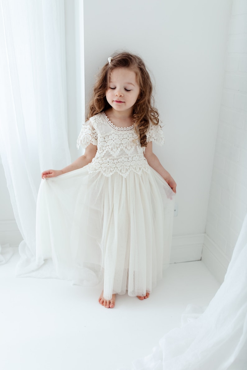 May include: A cream-colored girl's dress with a lace bodice and a flowing tulle skirt. The dress features short sleeves and a delicate lace neckline. The girl is standing in a bright, white-walled room, holding the skirt.