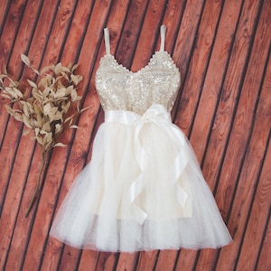 May include: A sleeveless dress featuring a gold sequined bodice with thin straps and a white tulle skirt. A wide white satin ribbon is tied in a bow at the waist. Beside the dress lies a decorative branch with dried, light brown leaves.