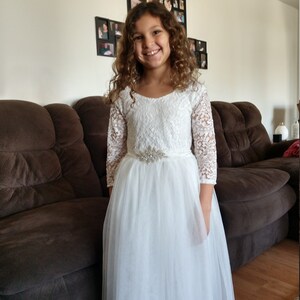 Puede incluir: Una ni&ntilde;a peque&ntilde;a con un vestido blanco de encaje con falda de tul y un cintur&oacute;n de cuentas plateadas. Est&aacute; sonriendo y mirando a la c&aacute;mara.