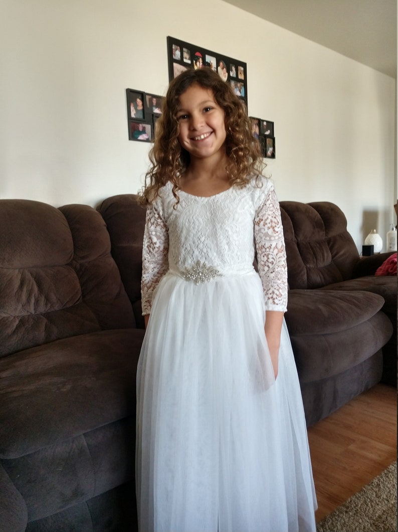 Puede incluir: Una ni&ntilde;a peque&ntilde;a con un vestido blanco de encaje con falda de tul y un cintur&oacute;n de cuentas plateadas. Est&aacute; sonriendo y mirando a la c&aacute;mara.