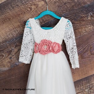 May include: A white lace dress with long sleeves and a tulle skirt. The dress has a coral flower sash with pearl accents.