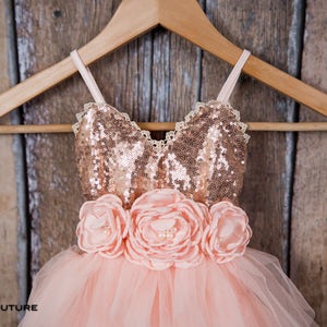 May include: A dress featuring a rose gold sequined bodice with thin straps and a lace trim neckline. The waist is adorned with three large peach-colored fabric flowers with pearl centers. The skirt is made of layered peach-colored tulle fabric.