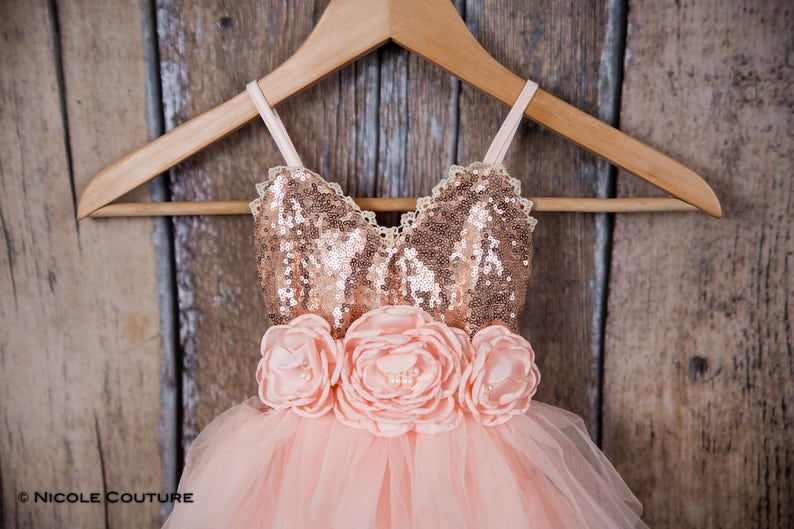 May include: A dress featuring a rose gold sequined bodice with thin straps and a lace trim neckline. The waist is adorned with three large peach-colored fabric flowers with pearl centers. The skirt is made of layered peach-colored tulle fabric.