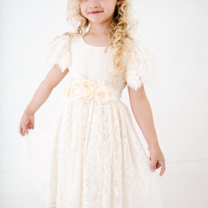 May include: An ivory lace dress featuring short flutter sleeves, a round neckline, and a waist adorned with three large fabric flowers. The skirt has a gathered, knee-length silhouette with a scalloped hemline, complemented by a floral headpiece.