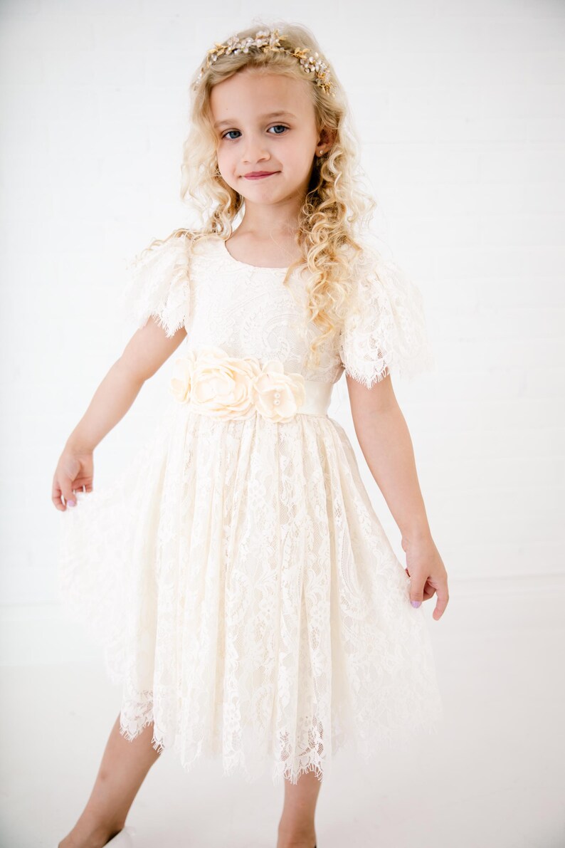 May include: An ivory lace dress featuring short flutter sleeves, a round neckline, and a waist adorned with three large fabric flowers. The skirt has a gathered, knee-length silhouette with a scalloped hemline, complemented by a floral headpiece.