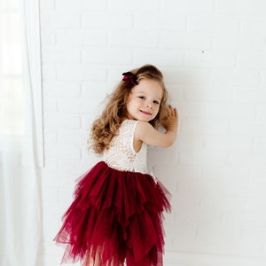 Puede incluir: Una ni&ntilde;a peque&ntilde;a con un vestido sin mangas con un corpi&ntilde;o de encaje blanco y una falda de tul burdeos en capas. Tiene el pelo largo y ondulado con un lazo burdeos. El fondo es una pared de ladrillos blancos.