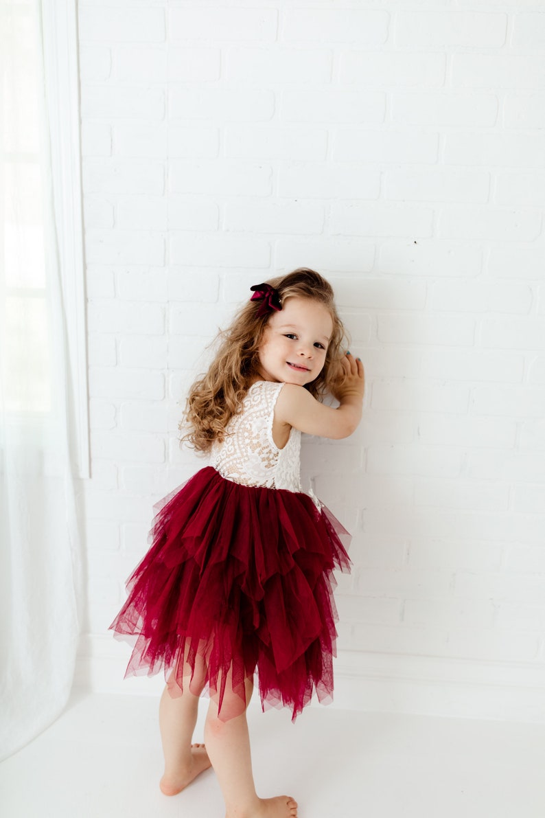 Puede incluir: Una ni&ntilde;a peque&ntilde;a con un vestido sin mangas con un corpi&ntilde;o de encaje blanco y una falda de tul burdeos en capas. Tiene el pelo largo y ondulado con un lazo burdeos. El fondo es una pared de ladrillos blancos.