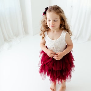Puede incluir: Una ni&ntilde;a peque&ntilde;a con un vestido de corpi&ntilde;o de encaje blanco y una falda de tul burdeos en capas. Tiene el pelo rubio rizado y un lazo burdeos. El vestido tiene un adorno de joya en la cintura.