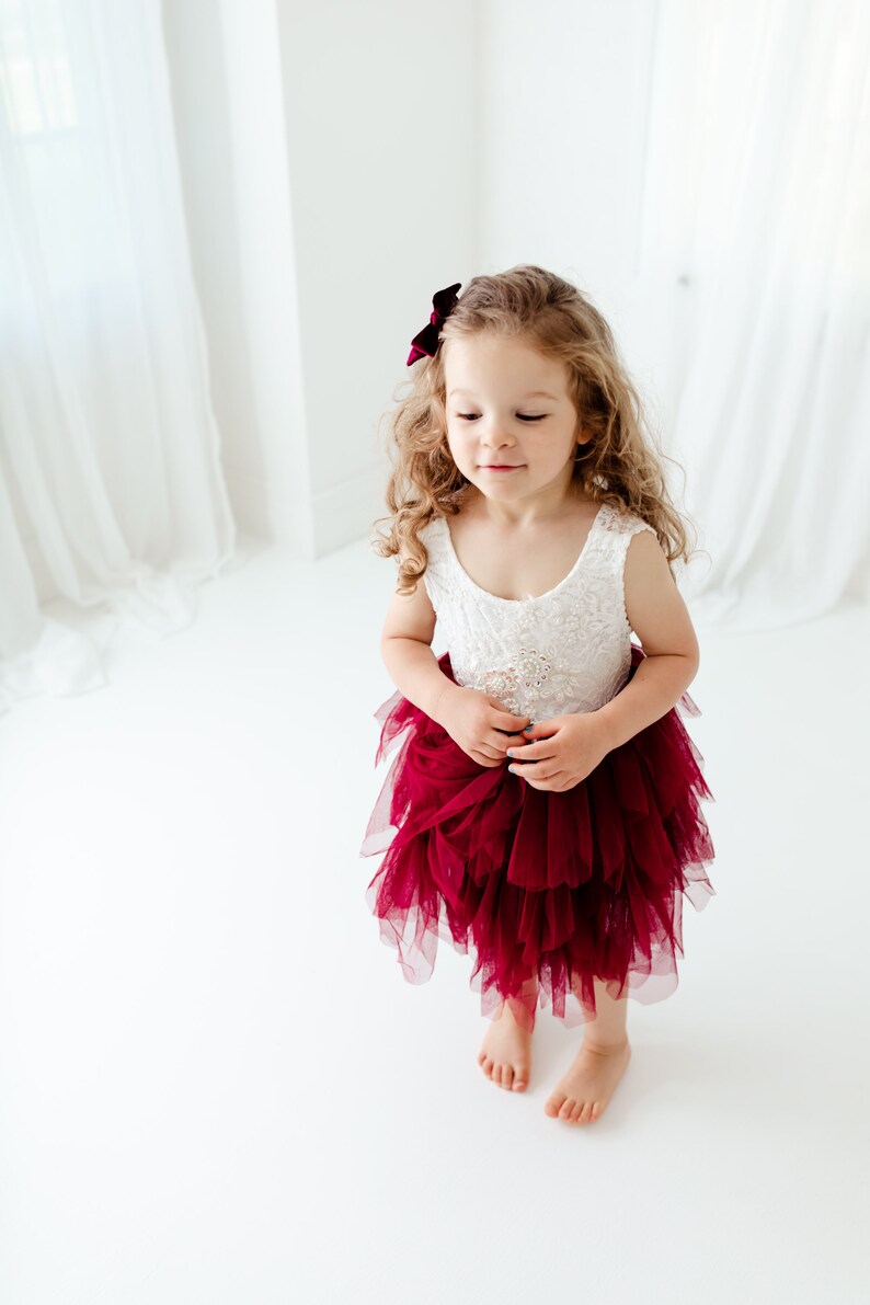 Puede incluir: Una ni&ntilde;a peque&ntilde;a con un vestido de corpi&ntilde;o de encaje blanco y una falda de tul burdeos en capas. Tiene el pelo rubio rizado y un lazo burdeos. El vestido tiene un adorno de joya en la cintura.