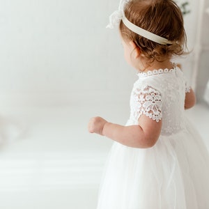 May include: A young child in a white lace dress with short sleeves and a tulle skirt. The dress features a high neckline and delicate lace detailing. Another child is in the background, also wearing a white dress. The room is bright and white.