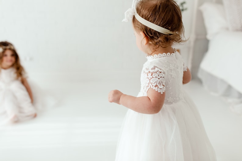 May include: A young child in a white lace dress with short sleeves and a tulle skirt. The dress features a high neckline and delicate lace detailing. Another child is in the background, also wearing a white dress. The room is bright and white.