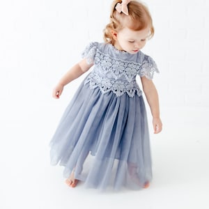 K&ouml;nnte beinhalten: Ein kleines M&auml;dchen tr&auml;gt ein hellblaues T&uuml;llkleid mit Spitzendetails. Das Kleid hat kurze &Auml;rmel und einen hohen Ausschnitt. Das M&auml;dchen tr&auml;gt eine rosa Schleife im Haar.