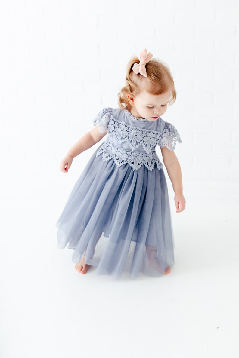 K&ouml;nnte beinhalten: Ein kleines M&auml;dchen tr&auml;gt ein hellblaues T&uuml;llkleid mit Spitzendetails. Das Kleid hat kurze &Auml;rmel und einen hohen Ausschnitt. Das M&auml;dchen tr&auml;gt eine rosa Schleife im Haar.