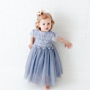 K&ouml;nnte beinhalten: Ein junges M&auml;dchen tr&auml;gt ein hellblaues T&uuml;llkleid mit einem Spitzenoberteil. Das Kleid hat einen gerafften Rock und kurze &Auml;rmel. Das M&auml;dchen steht vor einer wei&szlig;en Backsteinwand.