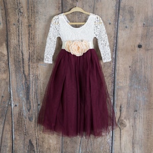 May include: A white lace long-sleeved dress with a burgundy tulle skirt and a peach flower sash.