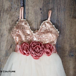 May include: A white tulle dress with a rose gold sequin bodice and a blush pink satin sash with three large blush pink fabric flowers.