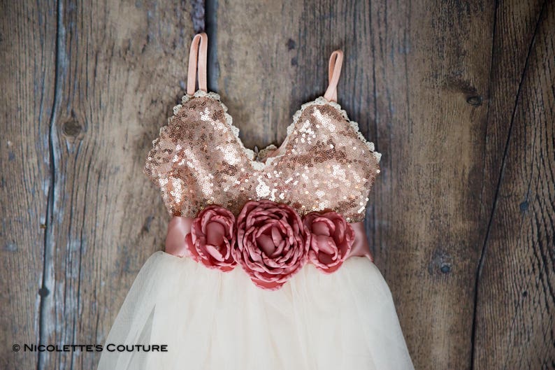 May include: A white tulle dress with a rose gold sequin bodice and a blush pink satin sash with three large blush pink fabric flowers.