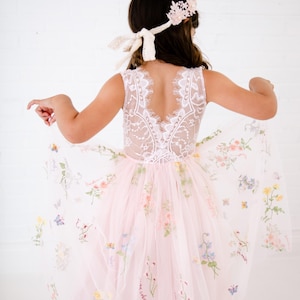 May include: A sleeveless dress with a white lace bodice featuring a deep V-back and a flowing pale pink tulle skirt adorned with colourful embroidered floral patterns and small butterflies. A matching floral headband with lace ties is worn.