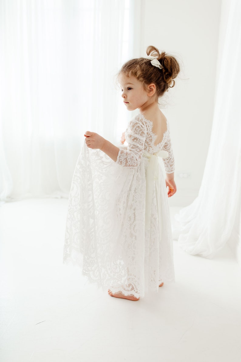 May include: A floor-length white lace dress featuring three-quarter length sleeves, a scalloped neckline, and a deep scoop back. The garment includes a solid white ribbon tied in a bow at the waist, with intricate floral lace patterns throughout.